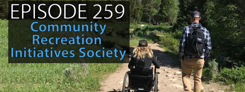 Image of a hiking trail with one able bodies person and one person in an adapted wheelchair hiking through a lush green set of trees.